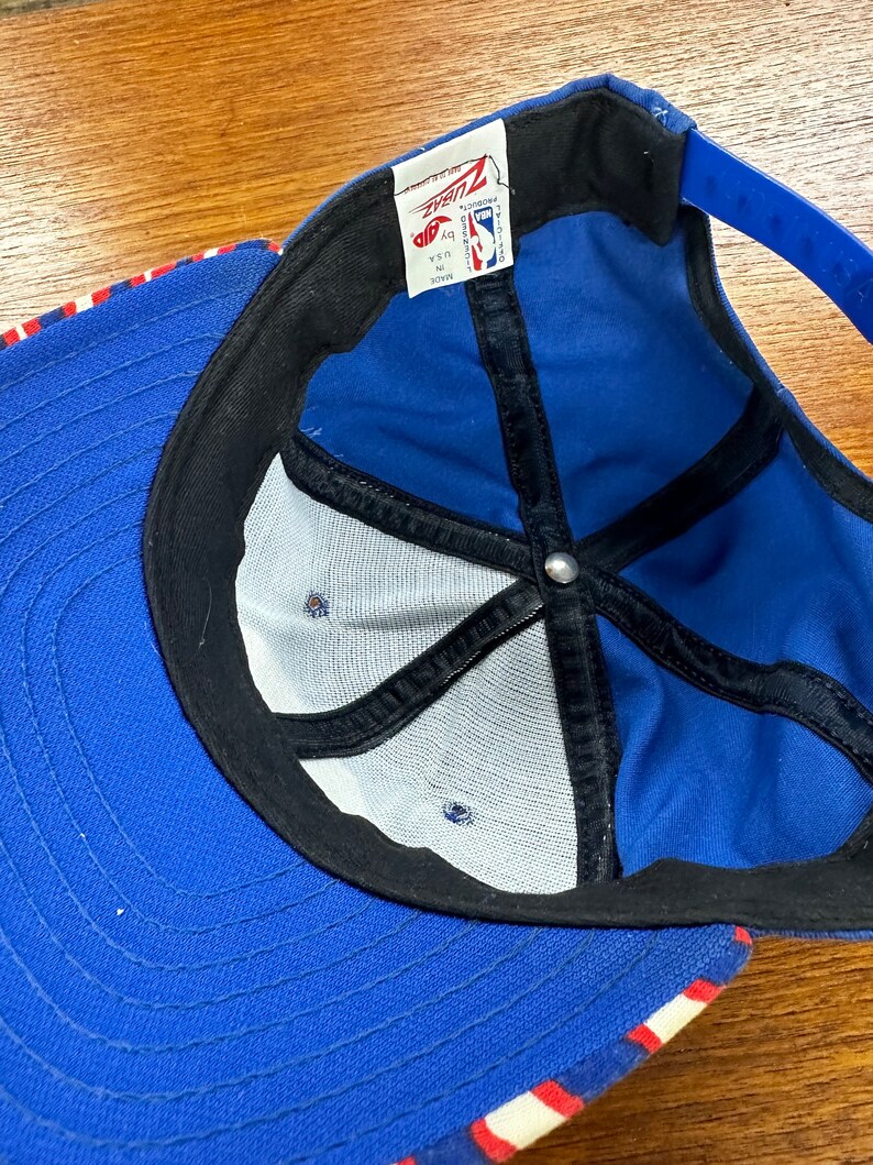 May include: Blue and white baseball cap with a red stripe. The cap has a mesh back and a black strap. The inside of the cap has a black label with the text "Ziaz" and "NBA Licensed Product".