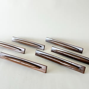 May include: A collection of seven rectangular, wooden incense holders. Each holder is a rich, dark brown color with a groove running down the center. The holders are arranged on a light beige surface.