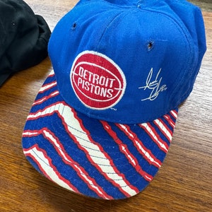 May include: A blue baseball cap with a red, white, and blue striped brim. The cap has a red circle with the words "Detroit Pistons" in white. The cap is signed with a black signature.