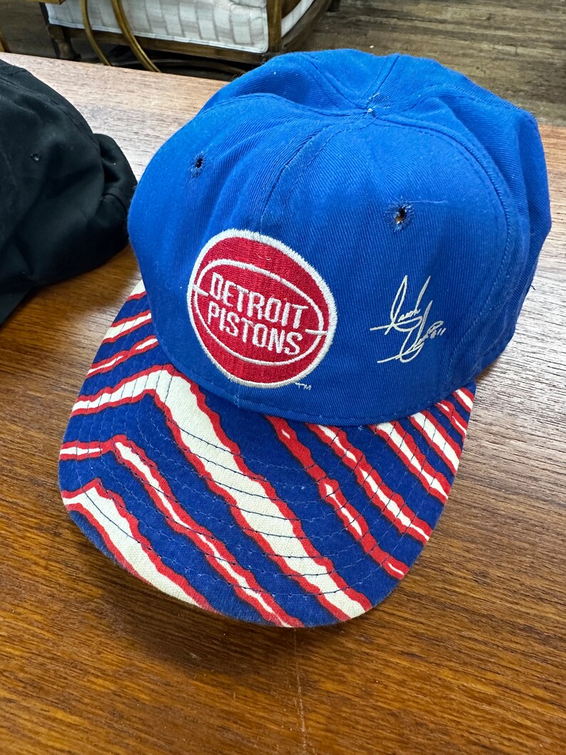 May include: A blue baseball cap with a red, white, and blue striped brim. The cap has a red circle with the words "Detroit Pistons" in white. The cap is signed with a black signature.