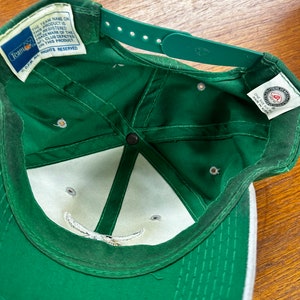 May include: Green baseball cap with a white brim and a white logo on the front. The cap has a velcro strap on the back and a tag that reads "Team Name on this product is the registered trademark of the NFL club depicted on this product. DPM All Rights Reserved."