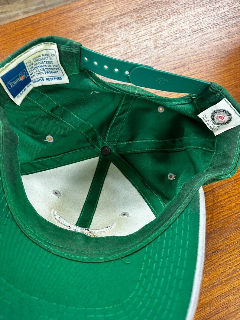 May include: Green baseball cap with a white brim and a white logo on the front. The cap has a velcro strap on the back and a tag that reads "Team Name on this product is the registered trademark of the NFL club depicted on this product. DPM All Rights Reserved."