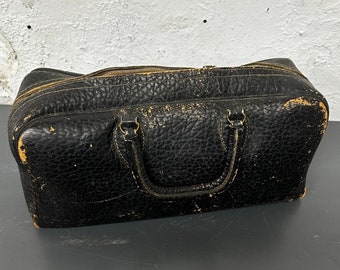 May include: A black leather travel bag with two handles and a zipper closure. The bag is worn and has a textured surface.
