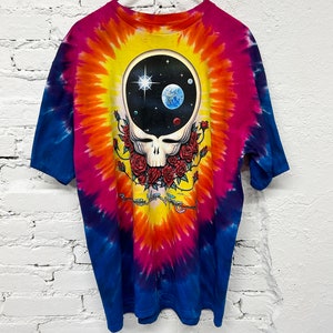 Puede incluir: Camiseta tie-dye con un patr&oacute;n de remolino colorido en tonos de azul, rosa, naranja y amarillo. La parte delantera de la camiseta presenta un gr&aacute;fico en blanco y negro de una calavera con rosas y una escena espacial dentro de la calavera.