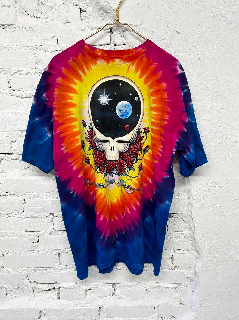 Puede incluir: Camiseta tie-dye con un patr&oacute;n de remolino colorido en tonos de azul, rosa, naranja y amarillo. La parte delantera de la camiseta presenta un gr&aacute;fico en blanco y negro de una calavera con rosas y una escena espacial dentro de la calavera.