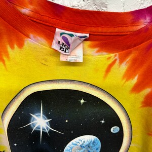 Puede incluir: Una camiseta tie-dye con un esquema de color amarillo, naranja y rojo. La camiseta presenta un gr&aacute;fico de una cabeza con un planeta en su interior. La etiqueta de la camiseta dice "Liquid Blue" y "XLarge".