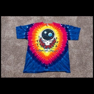 Puede incluir: Camiseta tie-dye con un dise&ntilde;o psicod&eacute;lico colorido de Grateful Dead Steal Your Face con una escena espacial dentro de la cabeza. La camiseta es una mezcla vibrante de azul, rosa, amarillo y naranja.