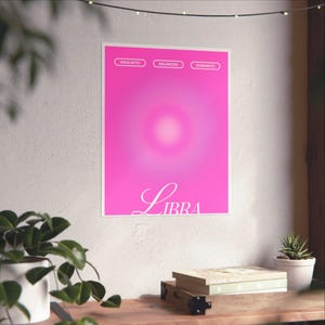 May include: A pink Libra poster with the words "Idealistic", "Balanced", and "Romantic" at the top. The word "Libra" is written in a cursive font at the bottom. The poster is displayed on a white wall.