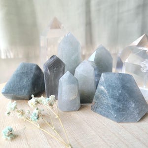 May include: An assortment of blue-green crystal points and geometric shapes. The crystals vary in size and shape, with some having a faceted appearance. Clear quartz crystals are also visible in the background. Dried flowers are in the foreground.