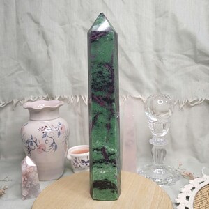 May include: A tall, polished zoisite crystal obelisk with a green and burgundy pattern. The crystal is displayed on a wooden surface, with a vase and other decorative items in the background. The obelisk has a pointed top.