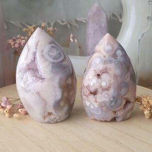 May include: Two teardrop-shaped agate stone sculptures. The stones feature a mix of white, lavender, and brown hues. The polished surfaces reveal intricate patterns and crystal formations. A light pink crystal tower is in the background.