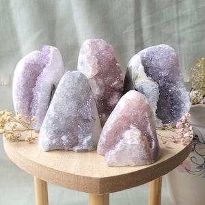 May include: A collection of amethyst crystal geodes in various shades of purple, lavender, and white. The geodes are displayed on a small, light-colored wooden table with a heart-shaped top. Dried flowers are placed around the geodes.