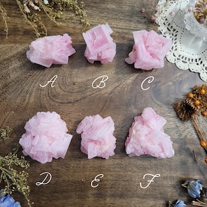 May include: Six pink crystal clusters are arranged on a wooden surface. The crystals vary in shape and size, with a soft, pastel pink hue. The crystals are labeled A through F.