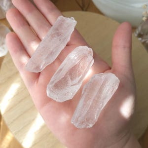May include: Three clear quartz crystals are held in a hand. The crystals are elongated and have a rough, natural texture. The crystals are translucent, allowing light to pass through them.