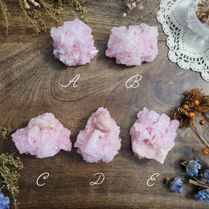 May include: Five pink calcite crystal clusters are displayed on a wooden surface. Each cluster has a unique shape and texture, with visible crystalline structures. The crystals are labeled A, B, C, D, and E. The crystals are a soft pink color.