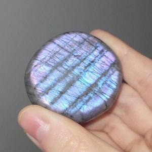 May include: A polished, oval-shaped labradorite gemstone. The stone displays a mix of gray, purple, and blue hues with iridescent flashes. The surface has a subtle, striped pattern. The gemstone is held in a hand.