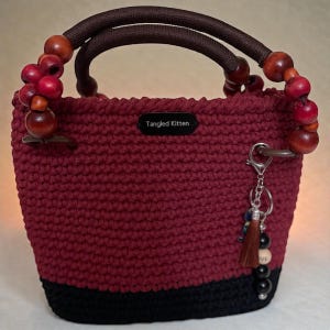 May include: A burgundy crochet handbag with a black base and brown handles. The handles are adorned with wooden beads. A small tag reads "Tangled Kitten". A keychain with a tassel and beads hangs from a metal ring.