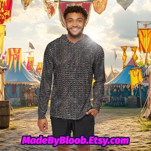 Puede incluir: Una persona con una camisa negra de manga larga con un patrón de cota de malla, de pie frente a un mercado medieval con tiendas y banderas.