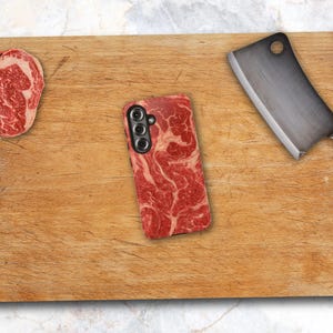 May include: A rectangular wooden cutting board featuring a raw steak slice on the left, a meat cleaver with a dark wooden handle on the right, and a smartphone case in the center with a red and white marbled raw meat pattern design.
