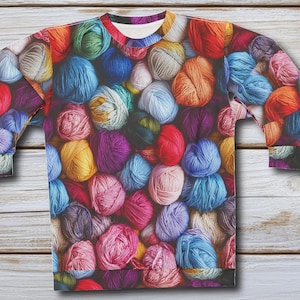 May include: A long-sleeved shirt with a colorful yarn print. The shirt is covered in balls of yarn in various colors, including blue, green, red, purple, and yellow.