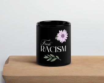 Può includere: Tazza in ceramica nera con la scritta "Fuck Racism" in corsivo bianco. Un fiore viola e foglie verdi sono presenti sulla tazza. La tazza è esposta su una superficie di legno.