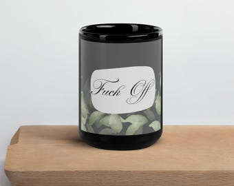 Può includere: Tazza in ceramica nera con le parole "Fuck Off" in elegante scrittura su un ovale bianco. La tazza presenta uno sfondo grigio scuro con un motivo a foglie verdi e poggia su una superficie di legno.