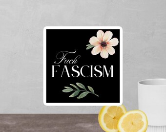 Può includere: Un adesivo quadrato con sfondo nero presenta il testo "Fuck FASCISM" in bianco, con un motivo floreale e di foglie. Un fiore color pesca e foglie verdi sono in alto e in basso. L'adesivo è su una superficie grigia con una tazza bianca e limoni a fette.