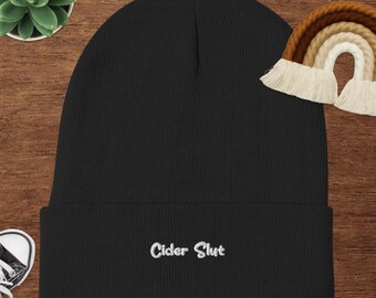 Può includere: Berretto nero con la scritta ricamata "Cider Slut" in bianco. Il berretto è realizzato in tessuto a maglia a coste. Un piccolo arcobaleno decorativo è sullo sfondo.