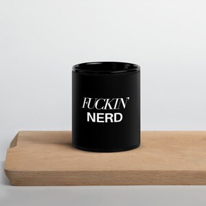 Puede incluir: Una taza de cerámica negra con las palabras "FUCKIN' NERD" impresas en blanco. La taza es cilíndrica y está sobre una tabla de cortar de madera marrón claro. El fondo es blanco liso.