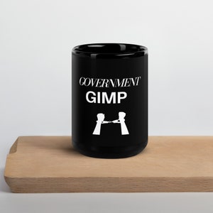 May include: Black ceramic mug with the words "GOVERNMENT GIMP" in white, along with a graphic of two figures holding handcuffs. The mug is on a wooden surface.