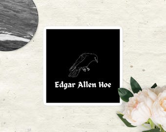 Può includere: Un adesivo quadrato con sfondo nero presenta un disegno a tratto bianco di un corvo. Il testo "Edgar Allen Hoe" è scritto in un carattere gotico sotto il corvo. L'adesivo è su una superficie strutturata bianco sporco.
