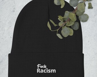 Può includere: Berretto nero con le parole "Fuck Racism" ricamate in bianco. Il berretto è realizzato in un materiale morbido e lavorato a maglia. Un rametto di foglie verdi è sullo sfondo.