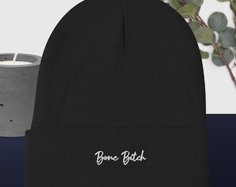 Può includere: Berretto nero con la scritta ricamata "Bone Bitch" in corsivo bianco. Il berretto è realizzato in morbido materiale a maglia. Lo sfondo include un portacandele in cemento e vegetazione.