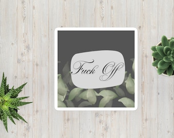 Può includere: Adesivo quadrato con la scritta "Fuck Off" in elegante corsivo. Il testo è su un ovale grigio chiaro su uno sfondo grigio scuro con foglie verdi. L'adesivo è su una superficie di legno.