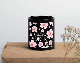 Può includere: Una tazza in ceramica nera con un motivo ripetuto di fiori di ciliegio rosa chiaro e il testo "Suck a DICK" stampato in un carattere serif bianco centrato sulla parte anteriore. La tazza ha una forma cilindrica e una finitura liscia.