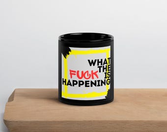 Può includere: Tazza in ceramica nera con la scritta "WHAT THE FUCK IS HAPPENING" in lettere rosse, gialle e bianche. Il testo è all'interno di un rettangolo giallo con accenti neri a pennello. La tazza è su una superficie di legno.