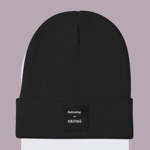 May include: A black beanie with a folded brim. A small, square patch is sewn on the front, displaying the text "Embracing my Cringe" in white. The beanie is made of a ribbed knit fabric.