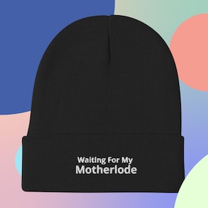 Può includere: Berretto nero con la scritta ricamata "Waiting For My Motherlode" in bianco. Il berretto è realizzato in un morbido materiale a maglia. Lo sfondo è un mix di blu, rosa e verde.