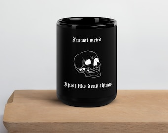 Può includere: Tazza in ceramica nera con grafica di teschio bianco e la scritta "I'm not weird, I just like dead things." La tazza cilindrica è appoggiata su una tavola di legno.