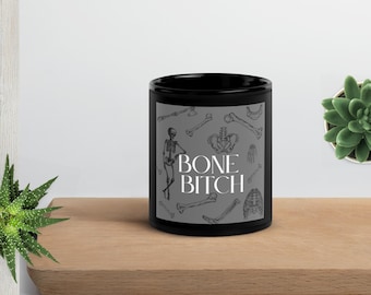 Può includere: Tazza in ceramica nera con sfondo grigio con varie illustrazioni scheletriche. La tazza ha il testo "BONE BITCH" in bianco. La tazza è appoggiata su una superficie di legno.