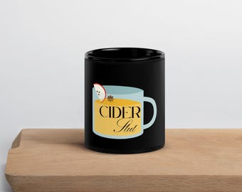Può includere: Tazza in ceramica nera con grafica di sidro, una fetta di mela e la scritta "CIDER Slut". La tazza è appoggiata su una superficie di legno.