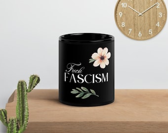 Può includere: Una tazza in ceramica nera con la scritta "Fuck Fascism" in corsivo bianco, accompagnata da un motivo floreale e un rametto di foglie. Un orologio in legno con lancette nere è sullo sfondo.