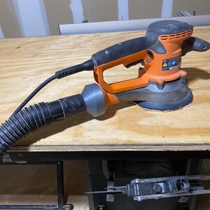 May include: An orange and black power sander with a dust collection hose attached. The sander is sitting on a wooden table.