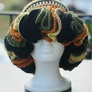 May include: A unique, handmade crochet hat in a variety of colors, including black, green, orange, and tan. The hat features a textured design with a brim and a decorative, curly accent. The hat is displayed on a white mannequin head.