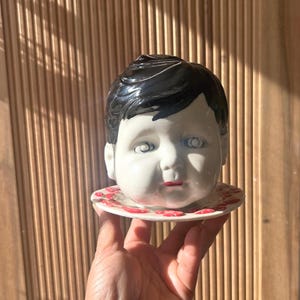 May include: A ceramic baby head figurine with black hair, white skin, and red lips. The head rests on a small, round plate decorated with red floral patterns. The figurine is held in a hand.