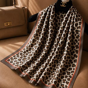 May include: A patterned silk scarf featuring a leopard print design in brown and black. The scarf has a cream background and a dark brown border with an orange trim. The scarf is displayed on a black hanger.