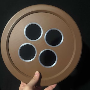 Op de afbeelding: Een grote, bruine knoop met vier zwarte cirkelvormige gaten en zilverkleurige randen. De knoop wordt door een hand tegen een donkere achtergrond gehouden. De knoop heeft een getextureerd oppervlak en een verhoogde buitenrand.