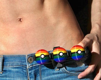 Op de afbeelding: Een riemaccessoire met drie bolvormige, regenboogkleurige ornamenten met een witte cirkel in het midden en een zwarte horizontale band, bevestigd aan een zwarte clip op de tailleband van een blauwe spijkerbroek.
