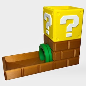 Op de afbeelding: Een dobbelsteentoren met een geel vraagtekenblok bovenop een bruine bakstenen basis met een groene pijpingang. De structuur heeft een rechthoekige opening aan de bovenkant en een schuin aflopend plateau aan de onderkant voor het rollen van dobbelstenen.
