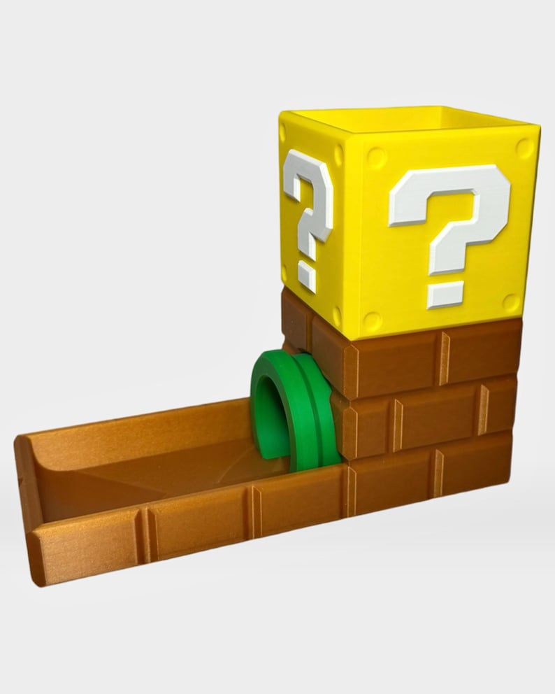 Op de afbeelding: Een dobbelsteentoren met een geel vraagtekenblok bovenop een bruine bakstenen basis met een groene pijpingang. De structuur heeft een rechthoekige opening aan de bovenkant en een schuin aflopend plateau aan de onderkant voor het rollen van dobbelstenen.